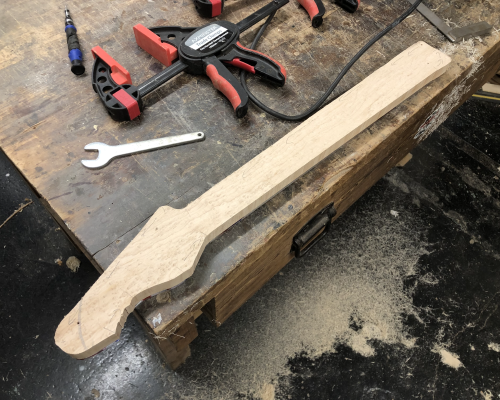 A work-in-progress guitar neck sits on the workbench, surrounded by sawdust on the floor.