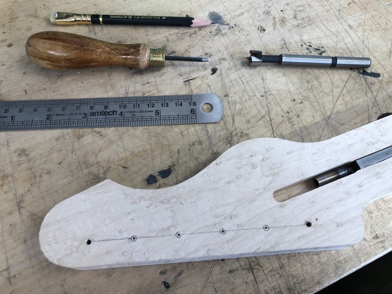 A close up on the headstock area of the neck in progress, showing the two screw holes from the template, and pencil markings from working out where the other 4 holes need to be, and little impressions from the bradawl to guide the drill bit. The drill bit sits ominously in the background.