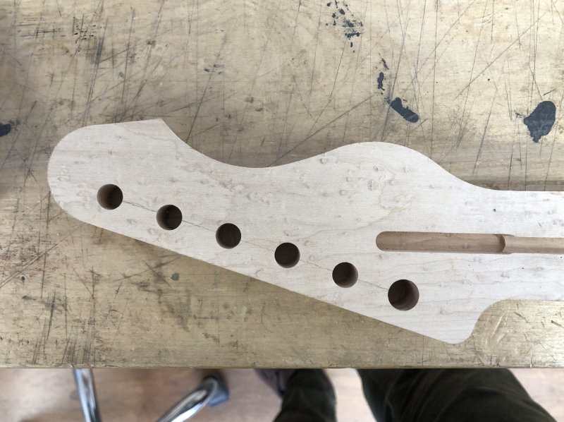 A close up of the headstock end of the neck-in-progress. There are 6 holes in the headstock in a neat line, 5 of which are the same size, and one of which is somewhat larger.