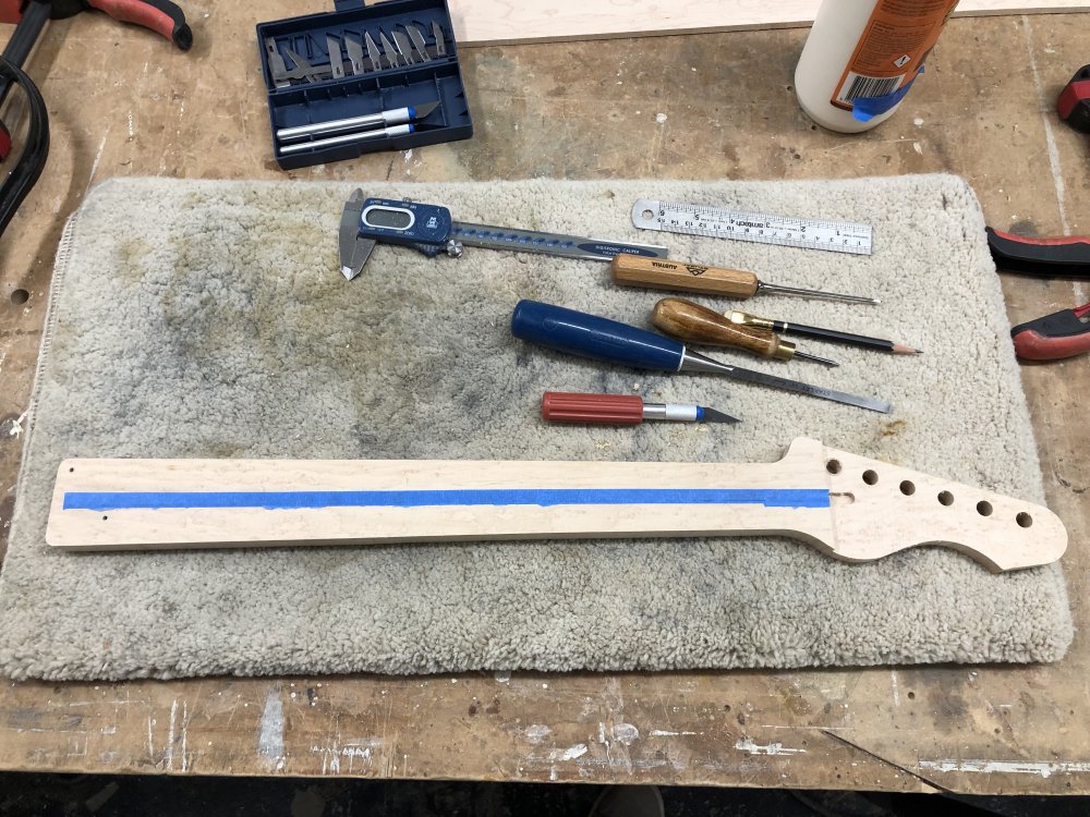 The neck sits on the workbench next to a mixture of chisels, measuring tools, a scaple, and a brawdal. Along the middle covering the truss-rod is a long thin strip of masking tape.