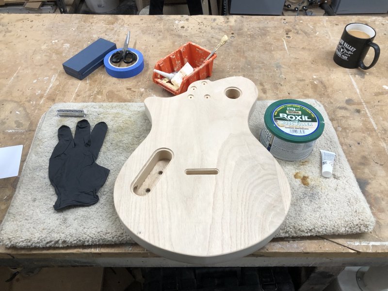 A guitar-body that is shaped and has all the holes in it, but is un-stained or finished sits face down on a square of carpet on a workbench. To one side is a tin of Roxil Wood Filler, to the other is a rubber glove, and then various implements for mixing and applying the filler.