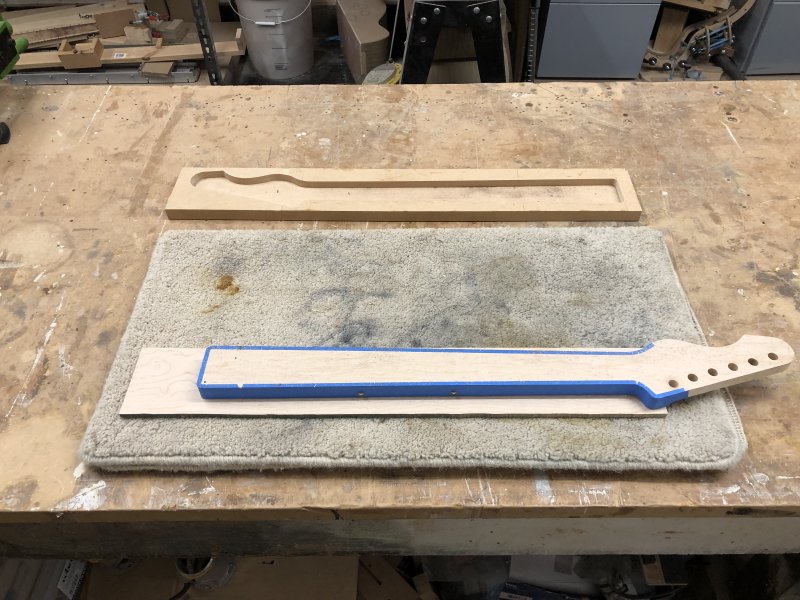 The neck sits on a carpet mat on top of a workbench, face down. you can see the fretboard material is still in a plank shape, yet to be trimmed flush with the rest of the neck. The main bit of the neck is covered in blue masking tape.