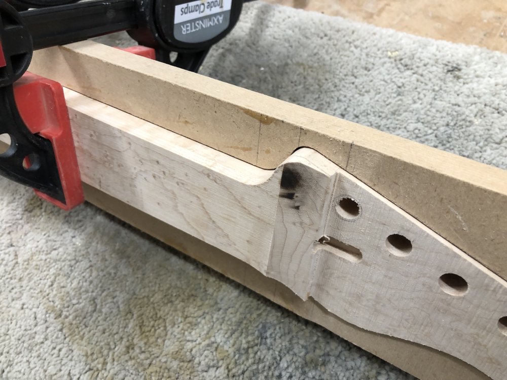 A close of up the in-progress headstock transition on the neck. You can see the step from the headstock to the fretboard has started to be turned into a curve, but on one side of that line the wood is has black marks on it. The neck is clamped into a jig that gives me a flat edge that is aligned with the center line of the neck for jobs like this.