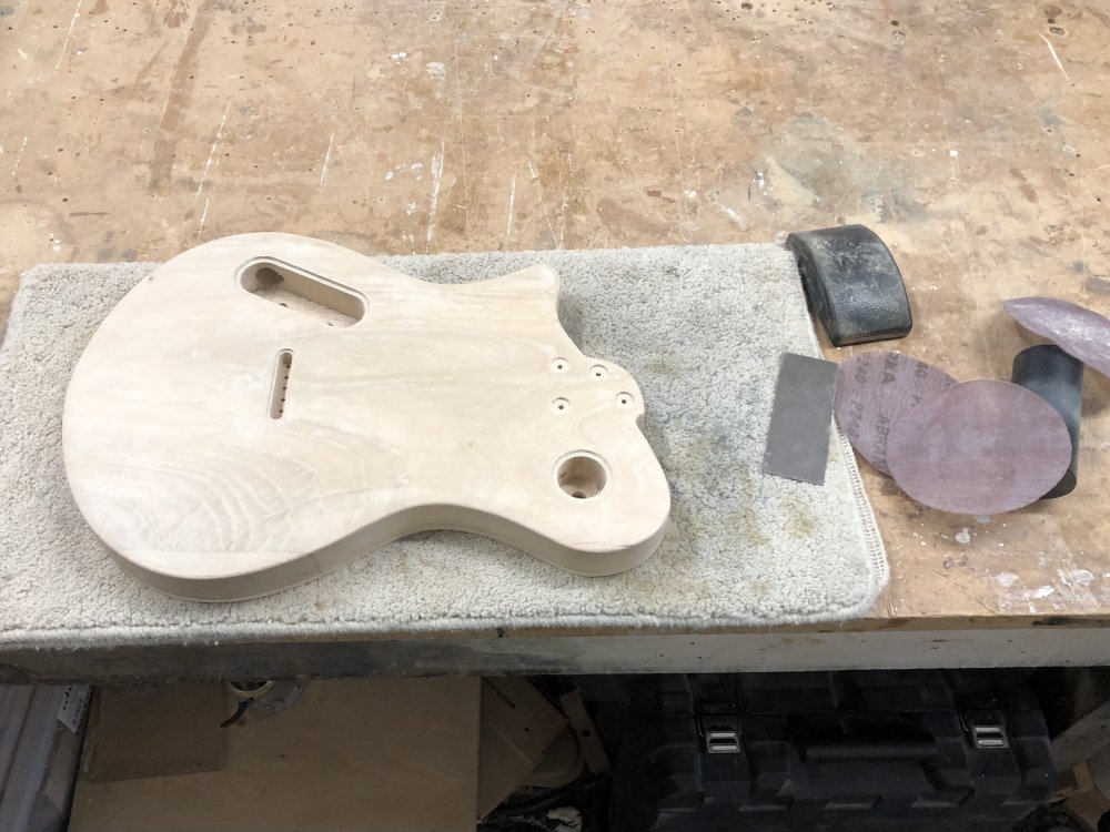 A guitar body sits face-down on a carpet mat on a workbench, next to which sits a metal scraper and some sanding-disks.
