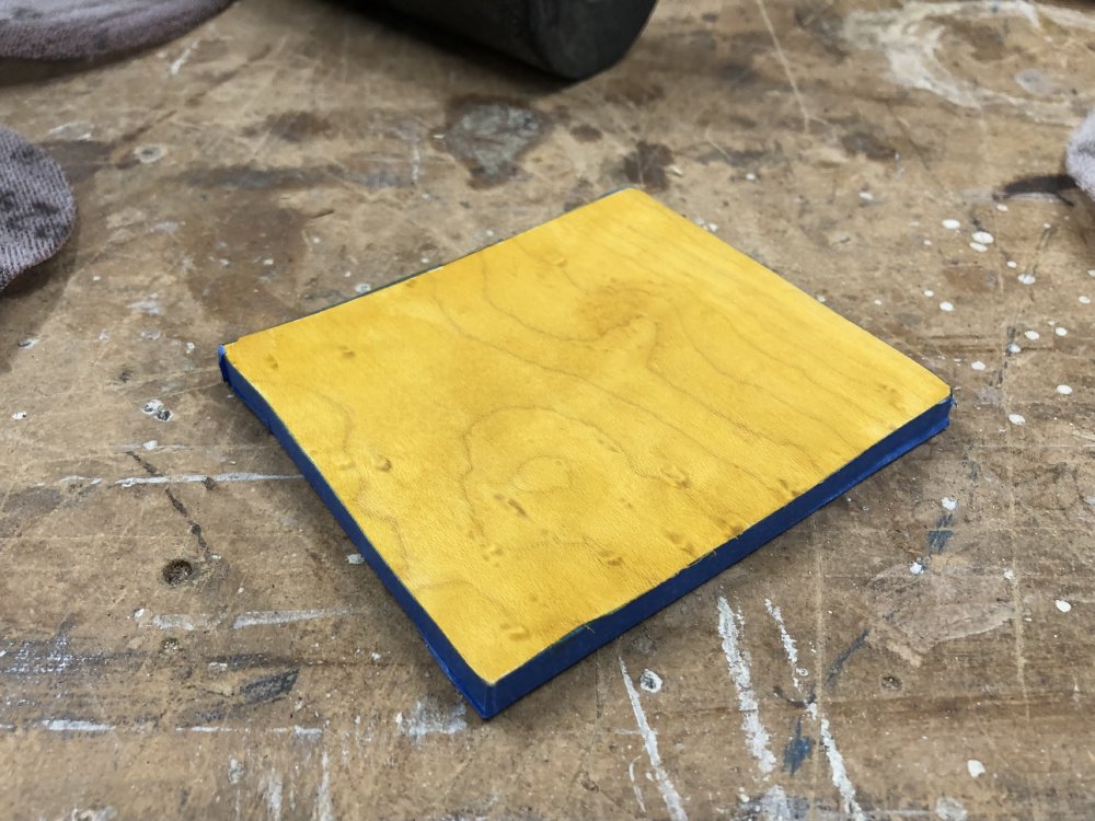 A square of birds-eye maple sits on the workbench. The front has been stained yellow, and all the sides are covered in blue masking tape.