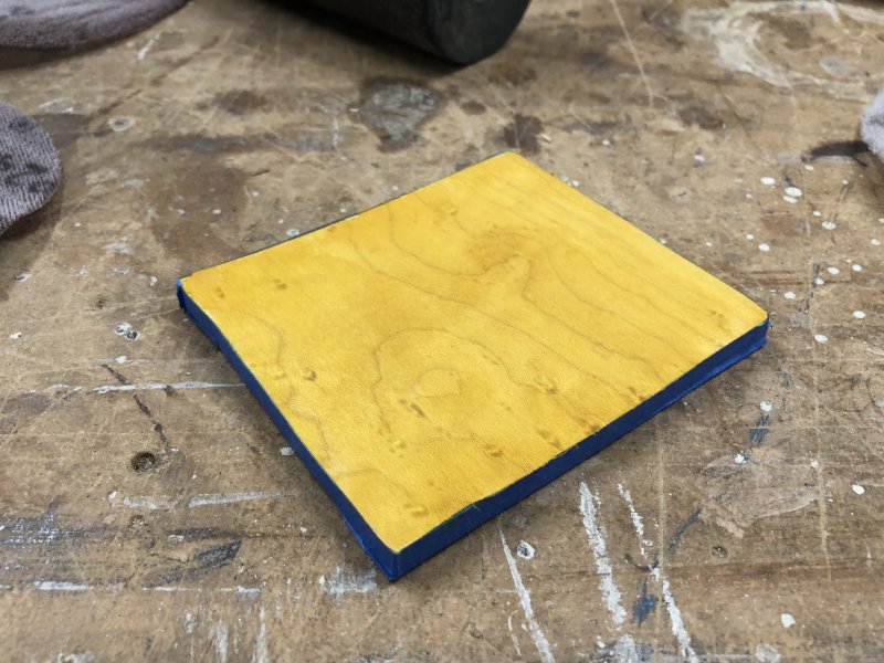 A square of birds-eye maple sits on the workbench. The front has been stained yellow, and all the sides are covered in blue masking tape.