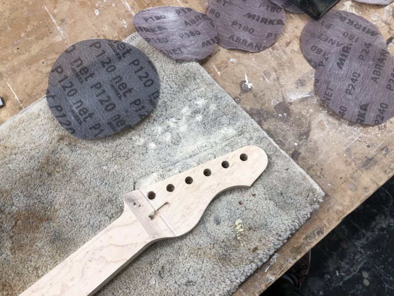 A picture of the headstock of the guitar on a carpet mat on a workbench, surrounded by wood-dust and sanding-disks.