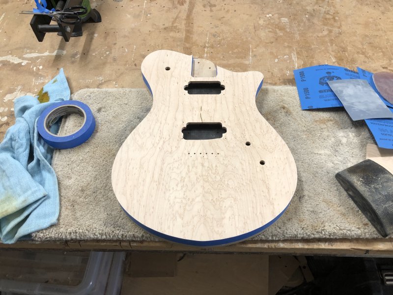 The guitar body sits on the workbench, face up, with blue masking tape running all the way around the top edge, including inside the neck pocket.