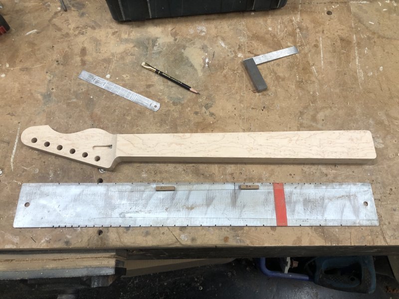 A guitar neck without fret slots cut sits on the workbench next to a long metal plate that has notches on both sides (each side is a different scale-length). There is also the outline of a neck drawn on the plate to help with positioning, and some wooden blocks glued to it to further help with alignment.