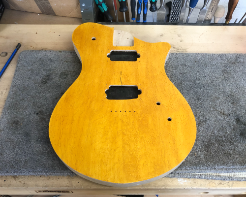 A guitar body sits on the workbench. It has a maple cap, and is stained a deep yellow colour.