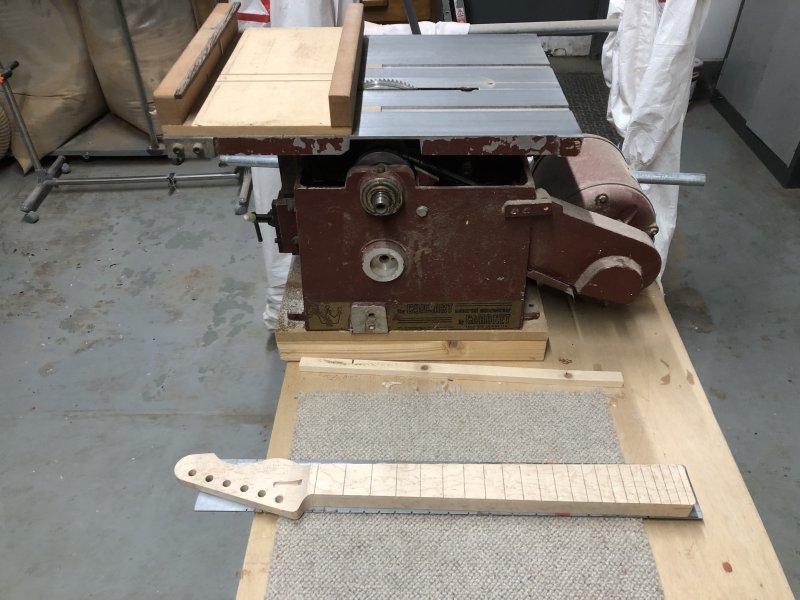 The neck, now with all 22 fret-slots cut, sits still stuck to the metal plate, besides an old looking small table-saw that has a wooden sled upon the table that lets you cut the fret-slots at a consistent depth.