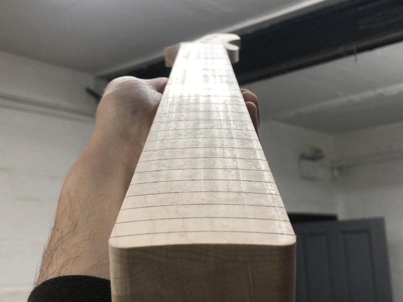 A view looking along the fretboard of the neck at a low angle from the heal to the headstock, pointed at an overhead light. In the reflections of the light on the fretboard you can see machine marks funning the length of the neck from where I cut the radius into the fretboard with a router.