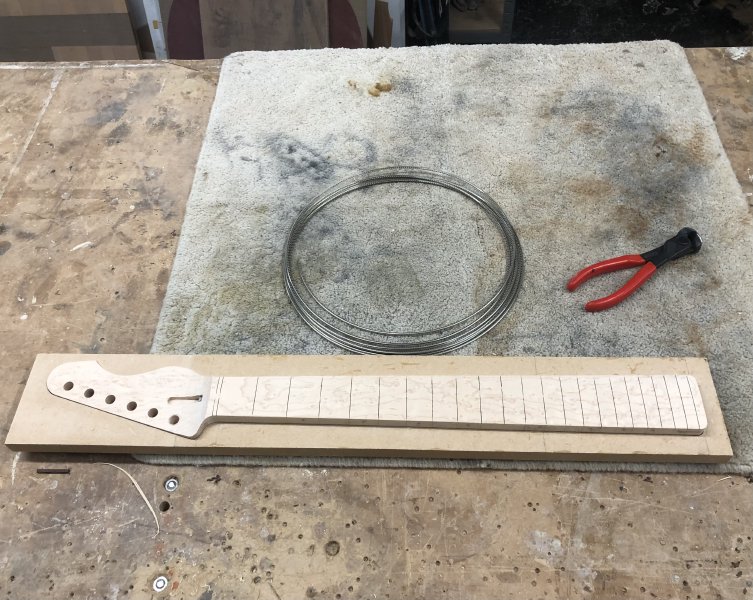 The neck sits on the workbench next to a long coil of fret-wire and a pair of metal cutting pliers.