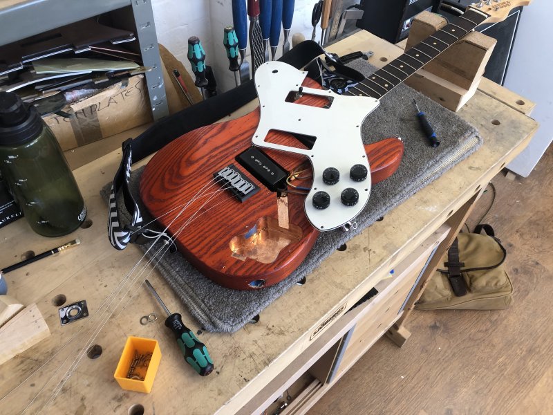 An orange coloured electric guitar sits on the workbench, with the pickguard removed and the routing for the electronics exposed underneath.