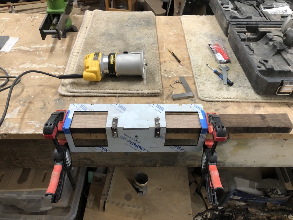 A plank of walnut wood is clamped to the edge of a workbench, with a laser-cut acrylic jig that sits on both the top and side of the wood (it's made from two parts stuck together). Behind it is the palm-router.
