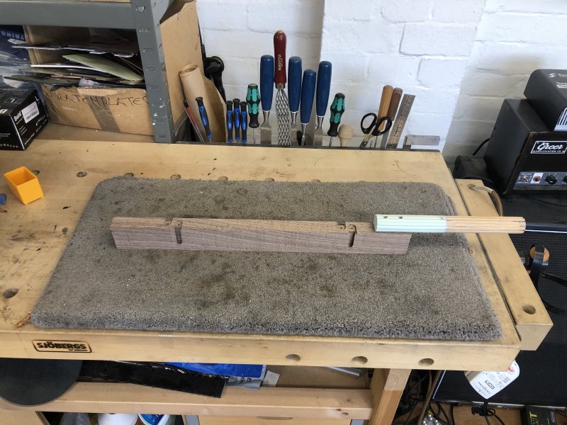 On another workbench, we see the plank now has slots cut into either side of it, and there is a rod of wood attached to it at one end for mounting it into the clamp in the spray booth.