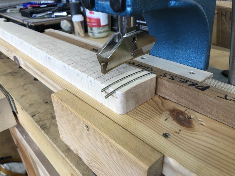The neck now sits under the arm of an arbour press. One fret has been pushed in, and another sits over the slot ready to join it.