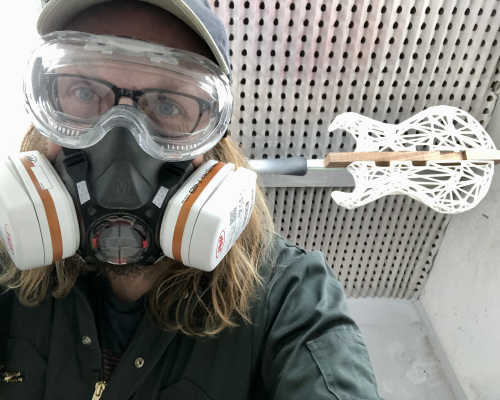 A selfie of me in the spray booth, wearing all my PPE (respirator, eye-protectors, goggles), and behind is the over-exposed 3D-printed body of a guitar ready for spraying with lacquer.