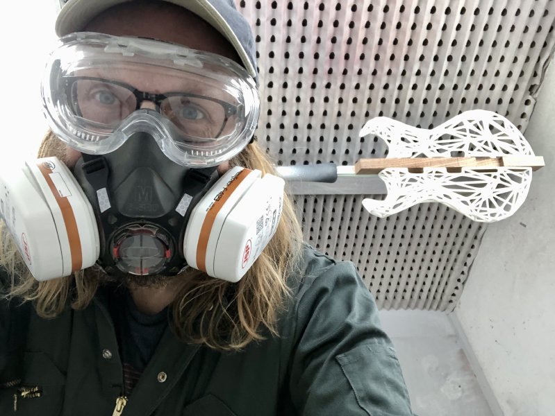 A selfie of me in the spray booth, wearing all my PPE (respirator, eye-protectors, goggles), and behind is the over-exposed 3D-printed body of a guitar ready for spraying with lacquer. My long hair however is escaping at all angles, and really should have been tied back.