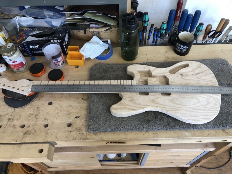 The in progress neck is matched with its in progress body on the workbench, and a long measuring stick is placed on top.