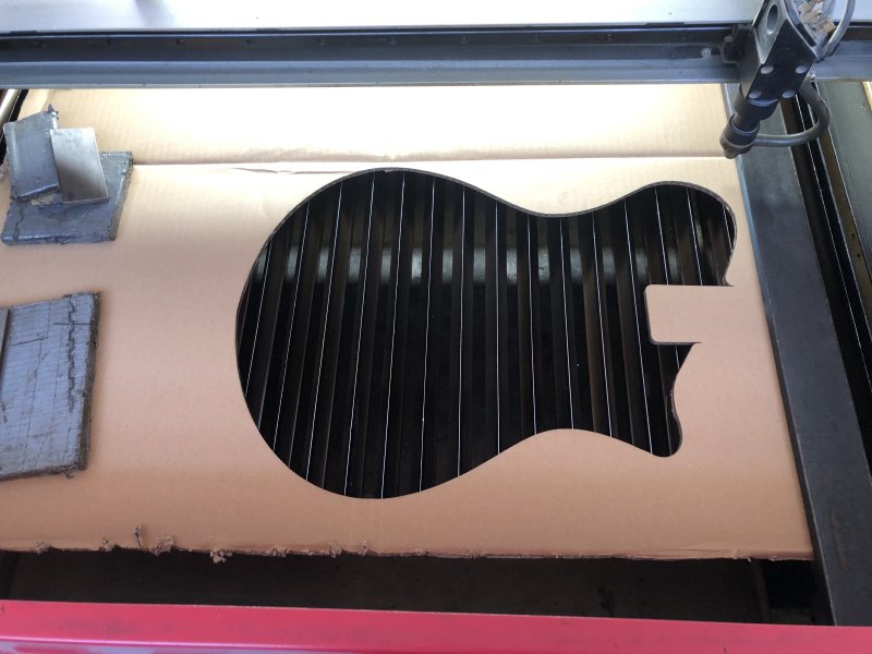 A sheet of cardboard sits on the bed of a laser cutter with a guitar body shaped hole in it.