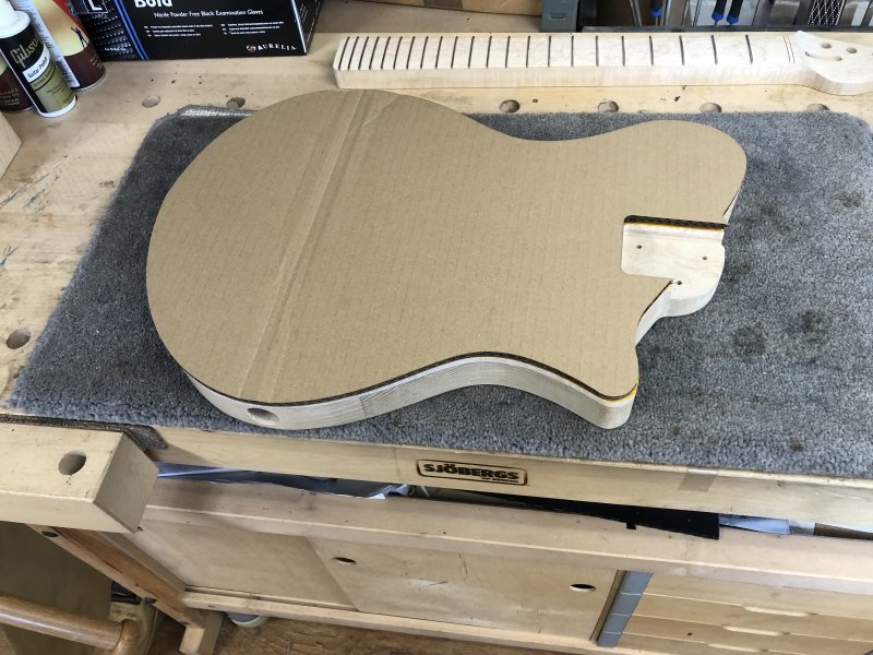 A guitar body sits on the workbench and on top the guitar body sits a perfectly matching cardboard cut-out of the body.