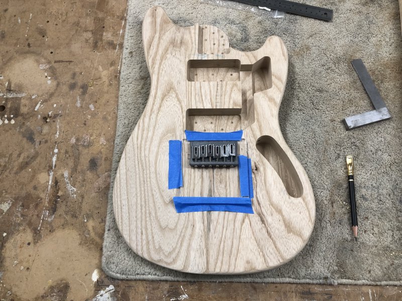 An in-progress guitar body sits on the workbench with a clear-acrylic template taped to where the bridge will mount, and atop the template sits a 3D-printed bridge with saddles.