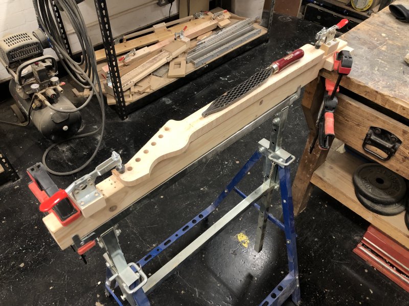 The neck sits fret-side down on a trestle, clamped at both ends, and upon the back sits the saw-toothed rasp ready to go to work.
