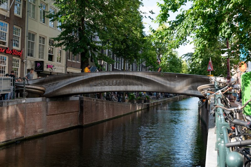 A photo of a metal looking foot-bridge over a canal surrounded by the stereotypical tall buildings of Amsterdam's old centre. The bridge has a wavy texture to it, making it clear this wasn't just made of straight rigid beams.