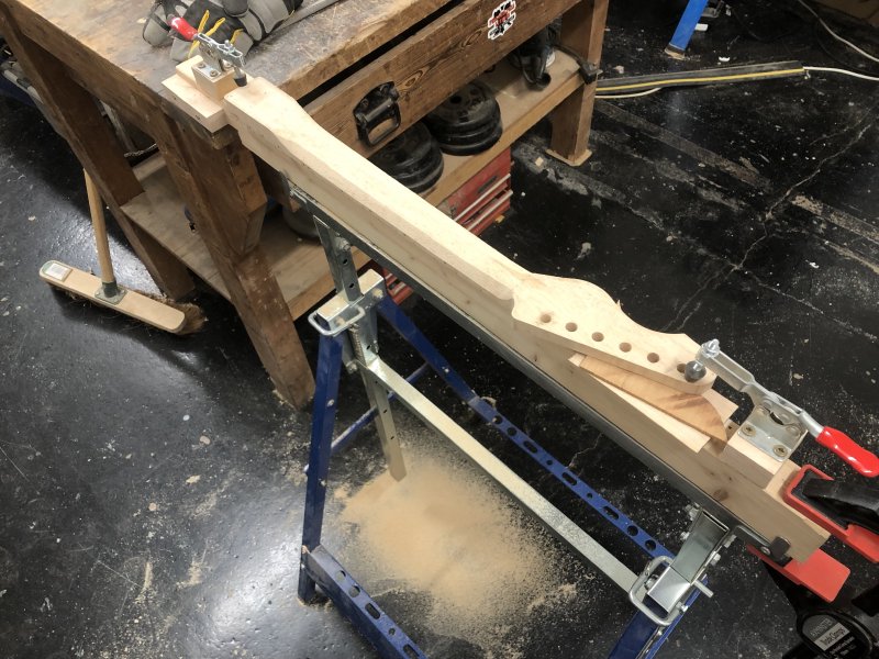 A view of an in-progress neck sat on a trestle, surrounded with wood-dust on the floor. You can see that the neck is still blockily shaped, with it mostly squared off from being cut from a plank with a template, but two angled facets have been carved along the length where you'll place your hand.