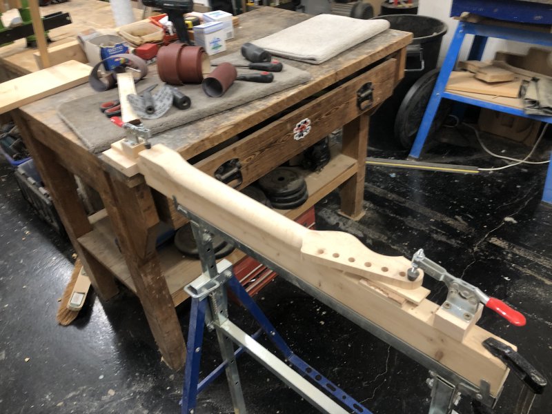 A blurry photo of the neck being carved as it sits on the trestle. Next to it are lots of spools of sand-paper.