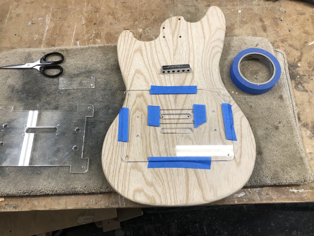 An unfinished, two-piece swamp-ash guitar body sits face down on the workbench atop a carpet tile, and on the back is  an acrylic template, stuck on with masking tape. Above the template you can see the ferrule block that will need to have a gap created for it, and to the sides you can see more bits of acrylic template.