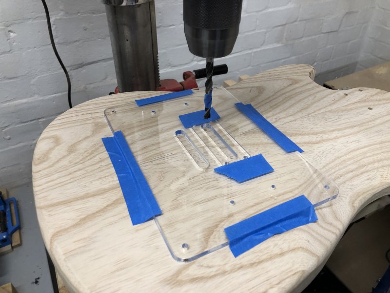 The guitar body with the router template stuck to it sits on the platform of a pillar drill, with a brad-point bit mounted in the chuck, and on the bit you can just about make a band of blue masking tape that'll tell me how far to drill in.
