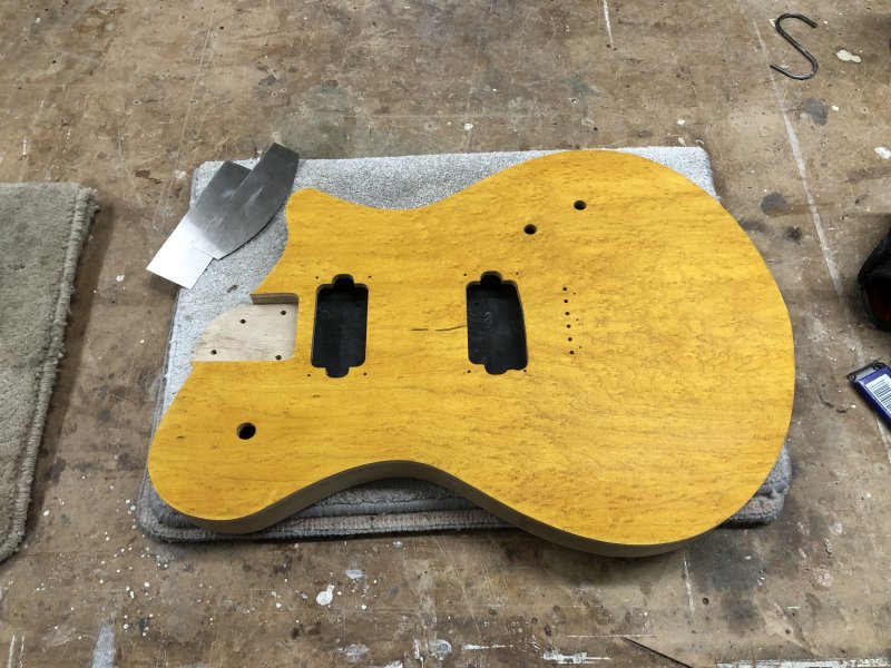 A different guitar body sits on the workbench upon a carpet tile. This one has the front, which is made from a figured birds-eye maple, stained a deep bright yellow colour, which contrasts with the black painted cavities for the pickups. Beside it sit two thin/flat pieces of metal that are wood scraping tools.