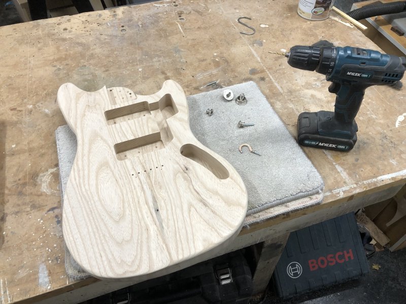 The guitar body sits on the workbench, next to which sit some strap buttons, a screw-in hook, and a handheld power-drill.
