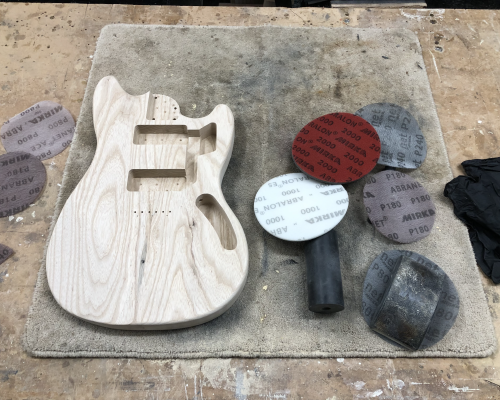 A shaped but unpainted guitar body sits on a workbench surrounded by sanding disk.