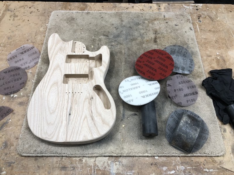 The shaped but unpainted guitar body sits on a workbench surrounded by sanding disk.