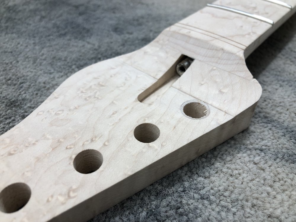 A close up photo of the headstock of the neck being worked on. Both the main neck body and fretboard are made from birds-eye maple. You can see just as the headstock face starts to curve up towards the fretboard is a small step in the wood, of less than a millimetre but still quite notable. You can also see in the smooth sweep from the headstock to the fretboard on the right hand side there is a small nick in the wood ruining the nice curve.