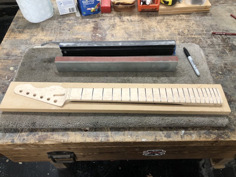 The neck sits on a workbench in a jig designed to stop it moving around, and beside it are two sanding bars about a foot long, and a black sharpie marker.