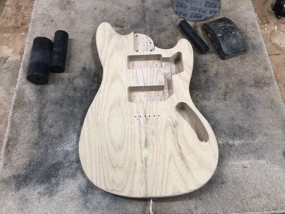 A guitar body sits on the workbench surrounded by sanding tools. The raw light wood of the body has a faint yellowy tinge to it from where the grain-filler has built up on the surface.