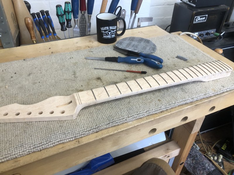 A guitar neck sits on the workbench, unfinished but fretted. Beside it sit a series of small files, a sanding pad, a razor blade, and a cup of tea.