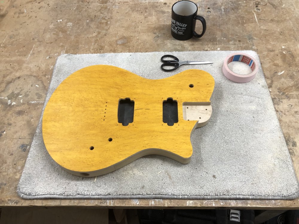 A guitar body sits on the workbench. The front face of the body has been stained yellow, letting you still see the patterning in the wood, but the sides are unfinished.