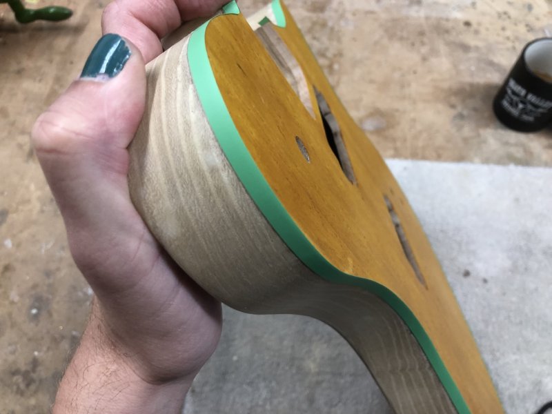 A close up on one side of the guitar body, where you can see a thin strip of green masking tape has been applied all along the edge towards the stained face.