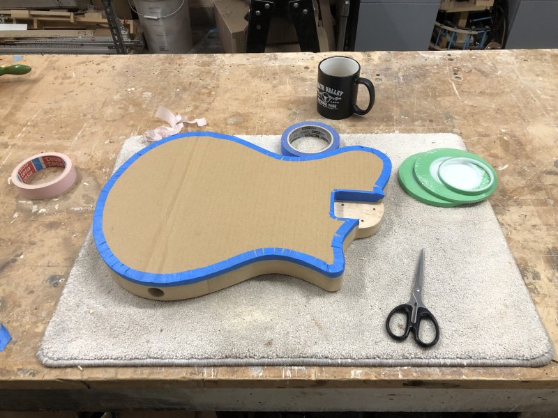 Similar to the first photo in this section, the guitar body sits on the workbench, but now it has a face covered in cardboard taken from a packaging box that has been cut to the same shape, and it's stuck on all edges with masking tape, sealing the front face in. Around the guitar body sit several rolls of masking tape, some scissors, and a cup of tea.