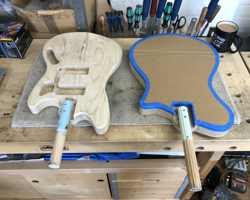 Two guitar bodies sit on the workbench, ready to go to the spray booth for painting. Both have a stick screwed into the neck pocket to be held in the clamp during spraying, and one has a cardboard mask on one side as only the rear will be painted.