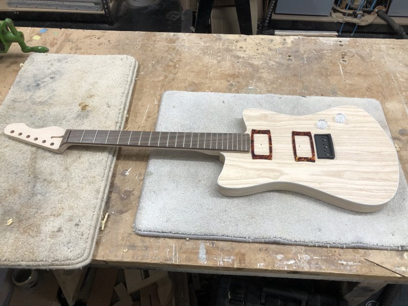 An in progress guitar sits on the workbench. The neck is mostly complete, though not yet oiled. The body is made from ash, and has white plastic binding on the upper edge. There are no holes in the front of the guitar for parts, and a bridge plate, pickup rings, and two knobs sit on the front to give an idea of where controls will be once complete.s
