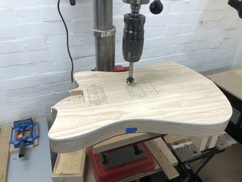 The guitar body sits once again on the plate of the pillar drill, but this time there is very obvious hatching in pencil where the pickup cavity will be, and a large 20mm wide forstner bit in the chuck. On the side of the guitar, about 1/3rd the way from the bottom, is a bit of blue masking tape.