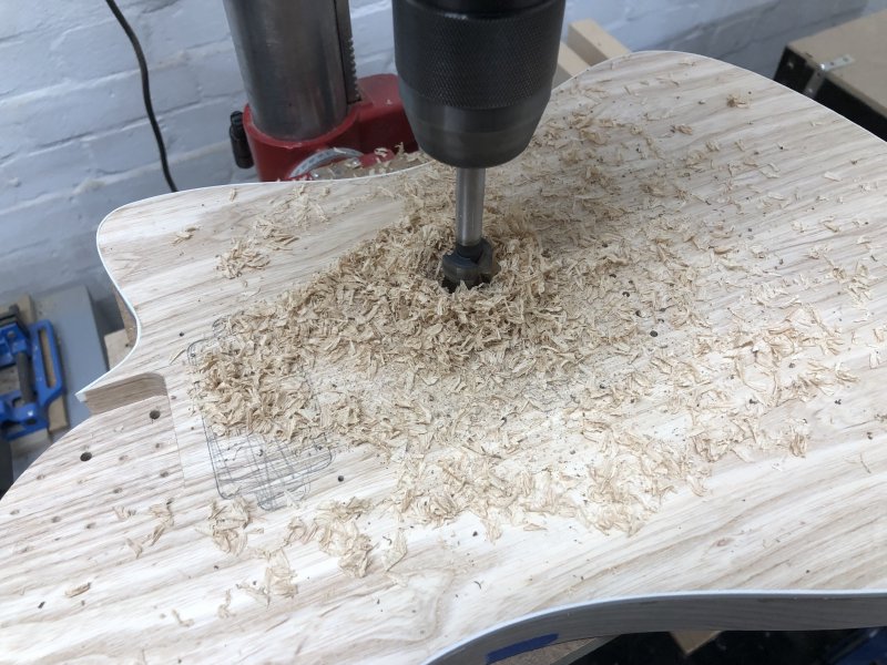 The same picture as before, with the guitar body on the drill press, but the top surface is covered with wood shavings and dust.