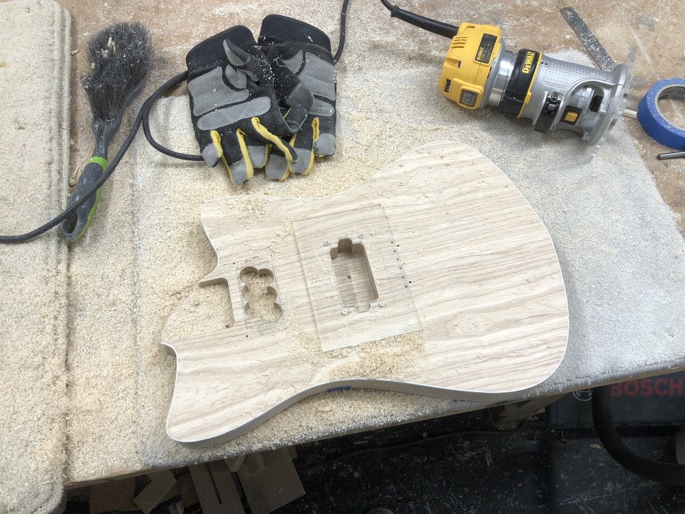 The guitar body is on the workbench, and on the more central pickup cavity is mounted the routing template again, and the cavity itself is now a nice smooth shape ready for a pickup, unlike the neck pickup cavity which is still just the six interlined holes. Beside it sits a dewalt palm router, and covering all the workbench is a lot of wood dust.