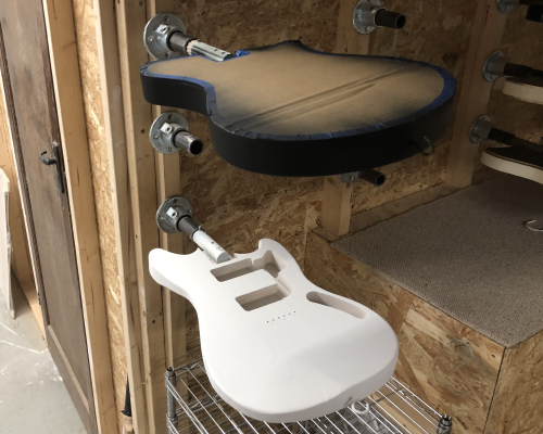 Two guitar bodies are mounted on a rack in the wall of the workshop, the lower one is painted in white primer, and the upper one has black primer around the sides, the top being covered with a cardboard screen.